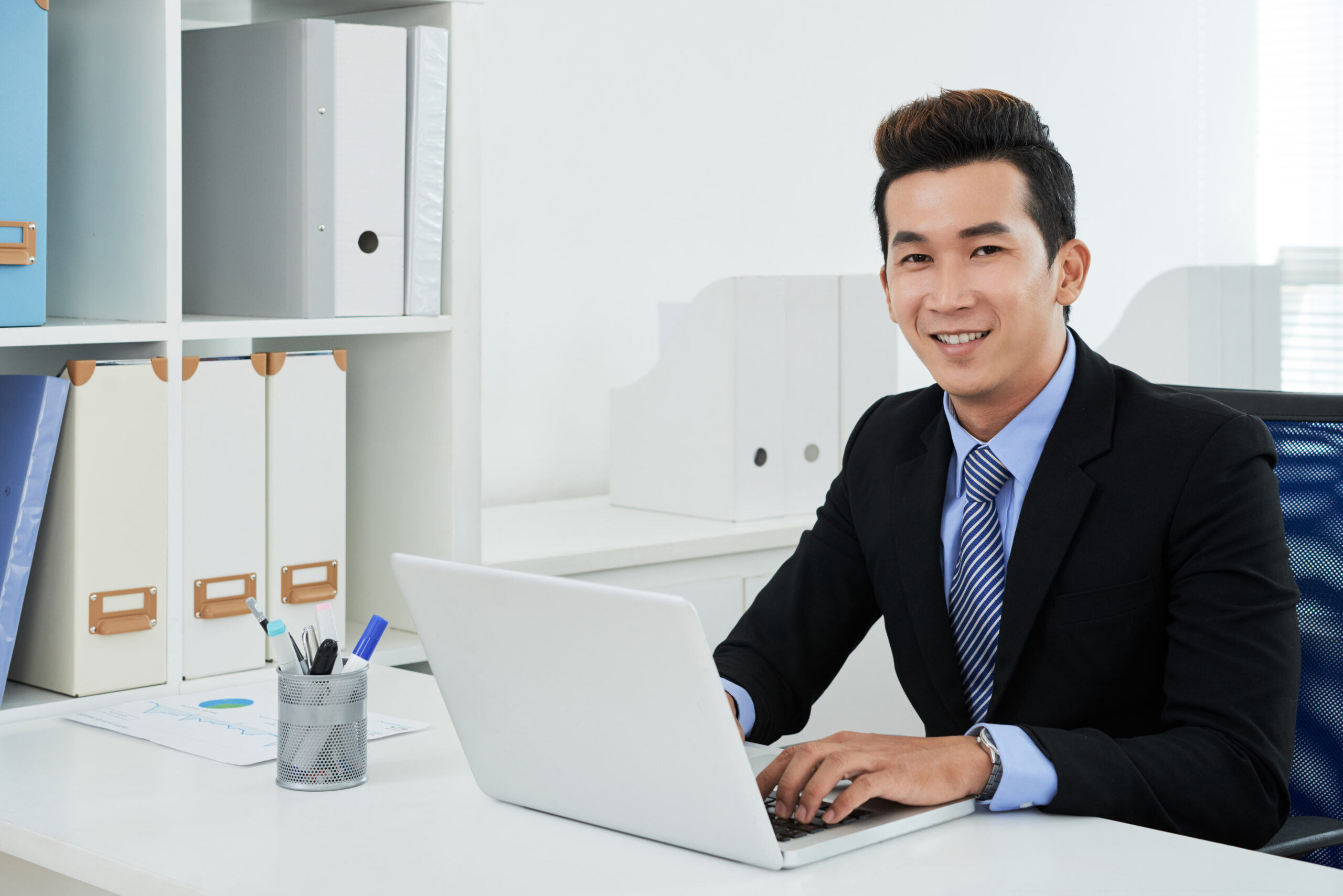 Handsome Asian white collar worker wearing suit sitting in front of modern laptop and posing for photography, modern office interior on background
